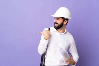 Miğferli genç bir mimar, elinde bir ürün sunmak için kenarı işaret eden izole edilmiş mor arka plan planları var.