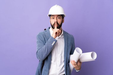 Miğferli genç bir mimar, izole edilmiş mor arkaplanının planlarını tutuyor. Parmağını ağzına koyduğunda sessizlik işareti gösteriyor.