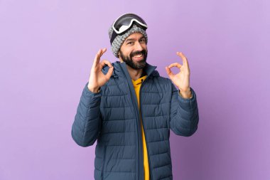 İzole edilmiş mor arka planda snowboard gözlüklü kayakçı iki eliyle tamam işareti gösteriyor.