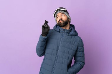 İzole edilmiş mor arka planda snowboard gözlüklü kayakçı adam parmaklarını çapraz yapıp en iyisini diliyor.