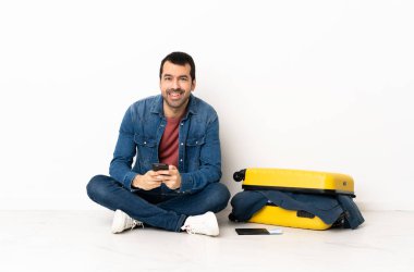 Kafkasyalı yakışıklı bir adam bavul dolusu kıyafetle içeride oturmuş cep telefonuyla mesaj yolluyor.