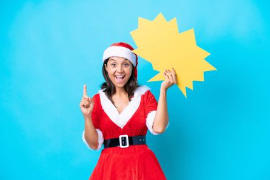 Anne Noel gibi giyinmiş genç İspanyol kadın mavi arka planda izole edilmiş. Elinde sürpriz bir ifadeyle boş bir konuşma balonu tutuyor.