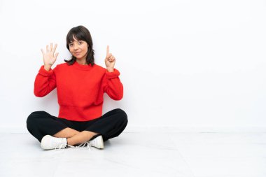 Genç melez kadın, beyaz arka planda izole bir şekilde yerde oturuyor. Altı parmaklı sayıyor.