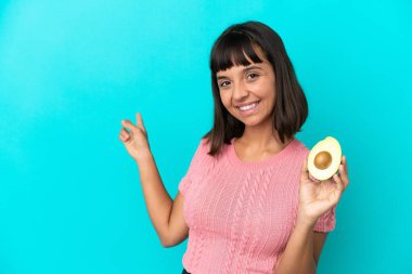 Mavi arka planda avakado tutan genç melez kadın geriye işaret ediyor.