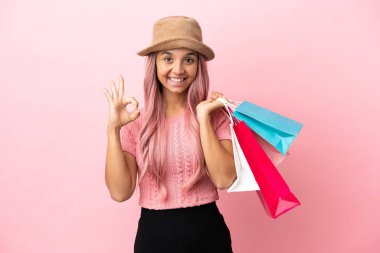 Pembe arka planda izole edilmiş alışveriş çantalı genç melez kadın parmaklarıyla tamam işareti gösteriyor.