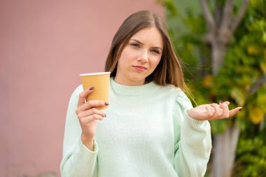 Genç, güzel, sarışın bir kadın açık havada kahve tutuyor. Omuzlarını kaldırırken şüpheyle hareket ediyor.