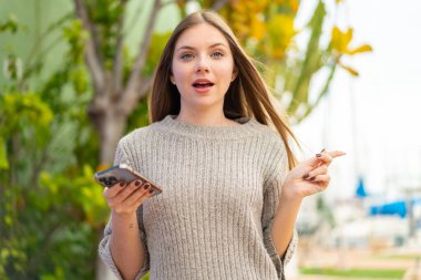 Açık havada cep telefonu kullanan genç, güzel sarışın kadın şaşırmış ve parmağıyla kenarı işaret ediyor.