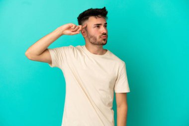 Kafkasyalı genç adam mavi arka planda izole edilmiş. Delilik jesti yapıyor. Parmağını kafaya koyuyor.