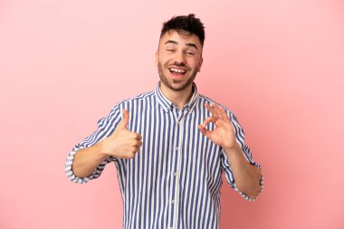 Pembe arka planda izole edilmiş genç beyaz bir adam tamam işareti ve baş parmak hareketi gösteriyor.