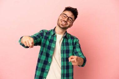 Pembe arka planda izole edilmiş genç beyaz adam mutlu bir ifadeyle ön tarafı işaret ediyor.