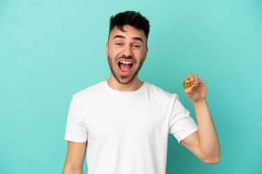 Mavi arka planda sürpriz bir yüz ifadesiyle izole edilmiş Bitcoin tutan genç adam.