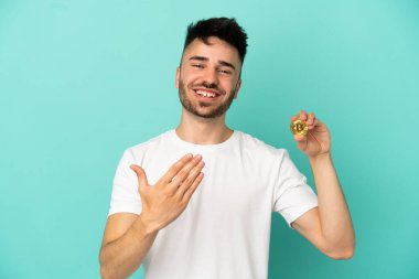 Elinde mavi arka planda izole edilmiş Bitcoin tutan genç bir adam, bizimle gelmesi için davet ediyor. Gelmene sevindim.