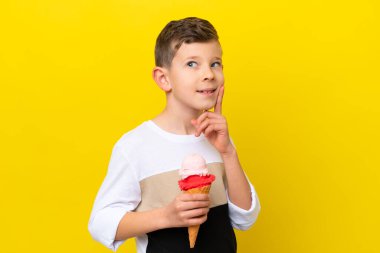 Kornet dondurması olan küçük beyaz bir çocuk sarı arka planda izole edilmiş bir şekilde yukarı bakarken bir fikir düşünüyor.