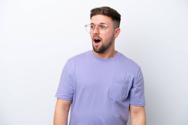 Beyaz arka planda izole edilmiş genç beyaz adam yan tarafa bakarken sürpriz jest yapıyor.
