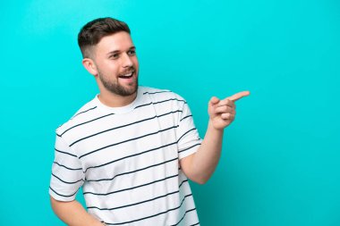 Mavi arka planda izole edilmiş genç beyaz bir adam parmağını yana doğru uzattı ve bir ürün sundu.