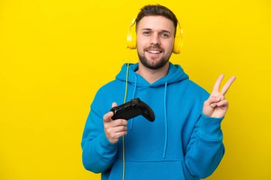 Sarı arka planda izole edilmiş bir bilgisayar oyunu kontrolörüyle oynayan genç beyaz adam gülümsüyor ve zafer işareti gösteriyor.