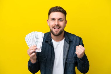 Kafkasyalı genç adam sarı arka planda izole edilmiş bir şekilde çok para alıyor ve galibiyeti kutluyor.