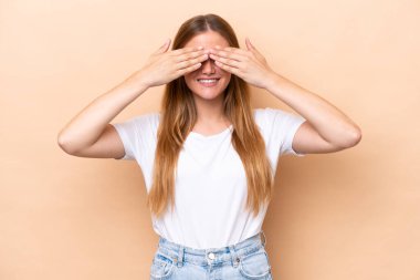 Beyaz tenli genç bir kadın bej arka planda izole edilmiş. Gözlerini elleriyle kapayıp gülümsüyor.