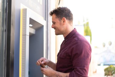 Açık havada ATM kullanan genç beyaz bir adam.