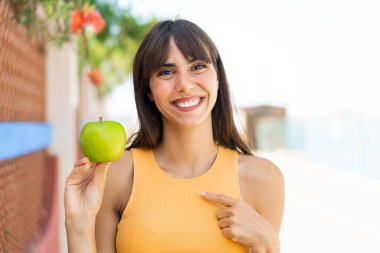 Açık havada elmalı genç bir kadın. Yüzünde sürpriz bir ifade var.