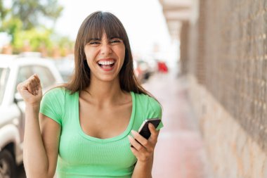 Dışarıda cep telefonu kullanan ve zafer jesti yapan genç bir kadın.