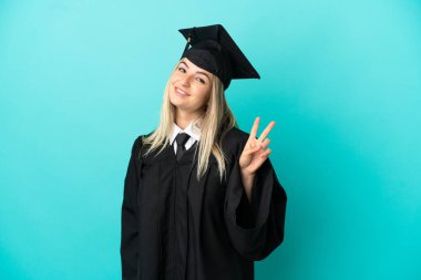 Genç üniversite mezunu mavi arka planda gülümseyerek ve zafer işareti göstererek.