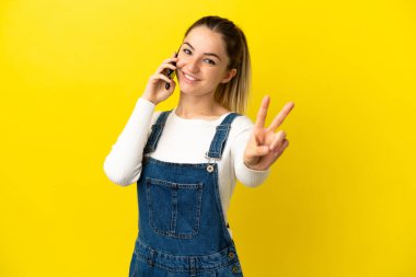 İzole edilmiş sarı arka planda cep telefonu kullanan genç bir kadın gülümsüyor ve zafer işareti gösteriyor.