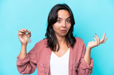 Latin kökenli genç bir kadın evinin anahtarlarını mavi arka planda tutarken ellerini kaldırırken şüpheye düşüyor.