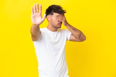 Sarı arka planda izole edilmiş beyaz, yakışıklı genç adam jesti durduruyor ve yüzünü kapatıyor.