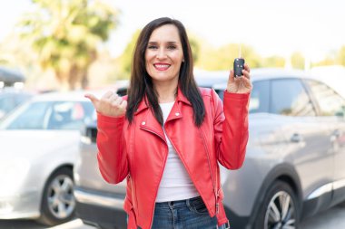 Orta yaşlı bir kadın dışarıda arabanın anahtarlarını tutuyor. Ürünü sunmak için yan tarafı işaret ediyor.