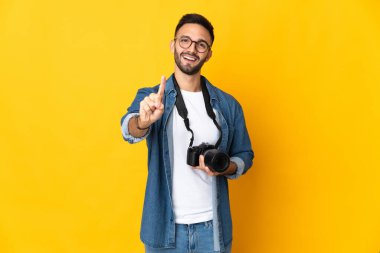 Sarı arka planda izole edilmiş genç fotoğrafçı kız parmağını kaldırıyor ve gösteriyor.