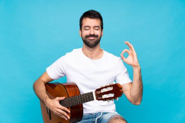 Gitarlı genç adam, izole edilmiş mavi arka planda zen pozunda.