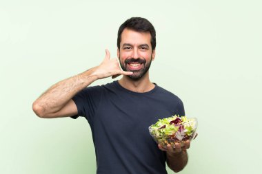 Soyutlanmış yeşil duvarın üstünde salatalı genç yakışıklı bir adam telefon jesti yapıyor. Bana geri dönüş işareti ver.