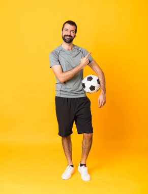 Futbol topuyla izole edilmiş sarı arka plan üzerinde tam boy bir adam fotoğrafı ve yan tarafı işaret ediyor.