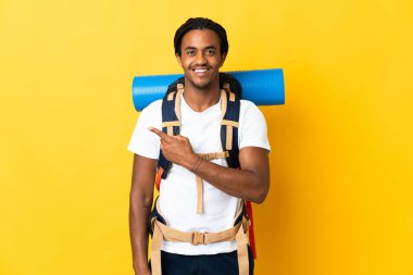 Sarı arka planda izole edilmiş büyük bir sırt çantalı genç dağcı bir ürünü sunmak için kenarı işaret ediyor.