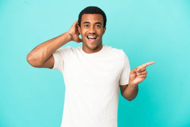 İzole edilmiş mavi arka plandaki Afro-Amerikalı yakışıklı adam şaşırdı ve parmağını yana uzattı.