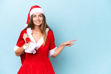 Anne Noel gibi giyinmiş genç beyaz bir kadın elinde Noel çuvalı tutuyordu. Mavi arka planda izole edilmişti. Şaşırmış ve parmağıyla yan tarafı gösteriyordu.