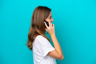 Mavi arka planda izole edilmiş genç beyaz bir kadın cep telefonuyla sohbet ediyor.
