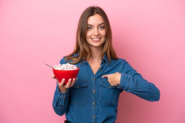 Young caucasian woman holding a bowl of cereals isolated on pink background with surprise facial expression