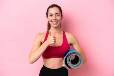 Genç beyaz kadın yoga derslerine giderken elinde pembe arka planda izole edilmiş bir minder tutarak baş parmak hareketi yapıyor.