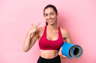 Genç beyaz kadın yoga derslerine giderken elinde pembe arka planda izole edilmiş bir minder tutarak baş parmak hareketi yapıyor.