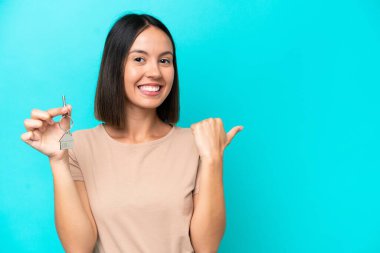 Elinde mavi arka planda izole edilmiş ev anahtarları olan genç bir kadın bir ürünü sunmak için kenarı işaret ediyor.