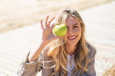 Genç, güzel sarışın Uruguaylı kadın açık havada elinde mutlu bir ifadeyle bir elma tutuyor.