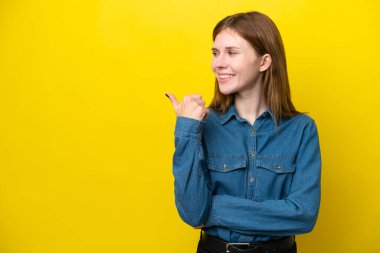 Sarı arka planda izole edilmiş genç bir İngiliz kadın bir ürünü sunmak için kenarı işaret ediyor.