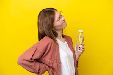 Sarı arka planda bir şişe suyu olan genç bir İngiliz kadın gayret gösterdiği için sırt ağrısından muzdarip.