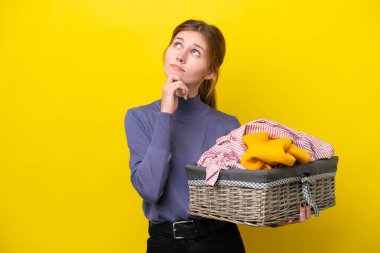 Young English woman holding a clothes basket isolated on yellow background and looking up