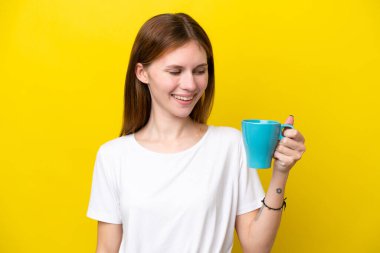 Güzel bir kadın, sarı duvar arkasında elinde sıcak bir fincan kahveyle mutlu bir şekilde gülümsüyor.