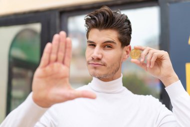 Açık havada elinde Bitcoin olan beyaz bir adam dur işareti yapıyor.