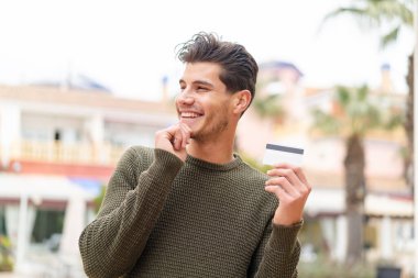 Açık havada kredi kartı tutan genç beyaz bir adam bir fikir üretiyor ve yan bakıyor.