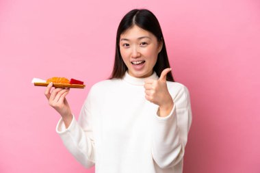 Genç Çinli bir kadın pembe arka planda saşimi tutuyor. Başparmakları havada. Çünkü iyi bir şey oldu.